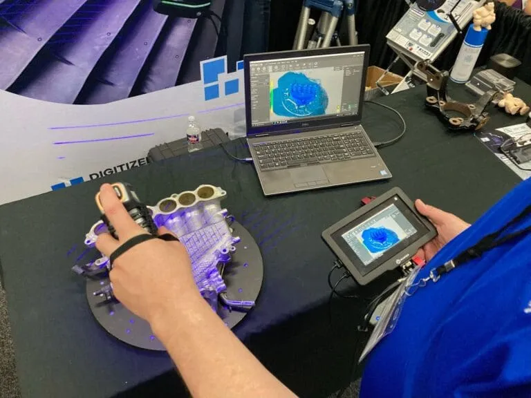 Handheld 3D scanner capturing an engine manifold on a turntable at a Digitize Designs trade show booth, with the scan on a tablet and laptop