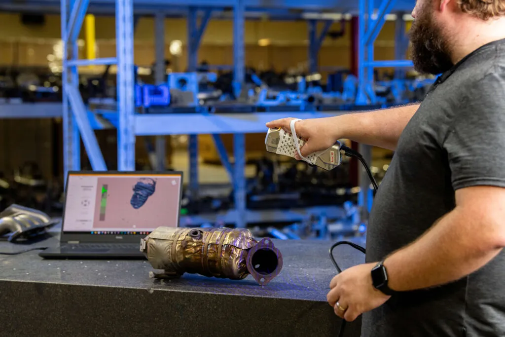 Technician 3D scanning an automotive exhaust part using the artec point 3d scanner, with a laptop displaying the live scan model in the background.