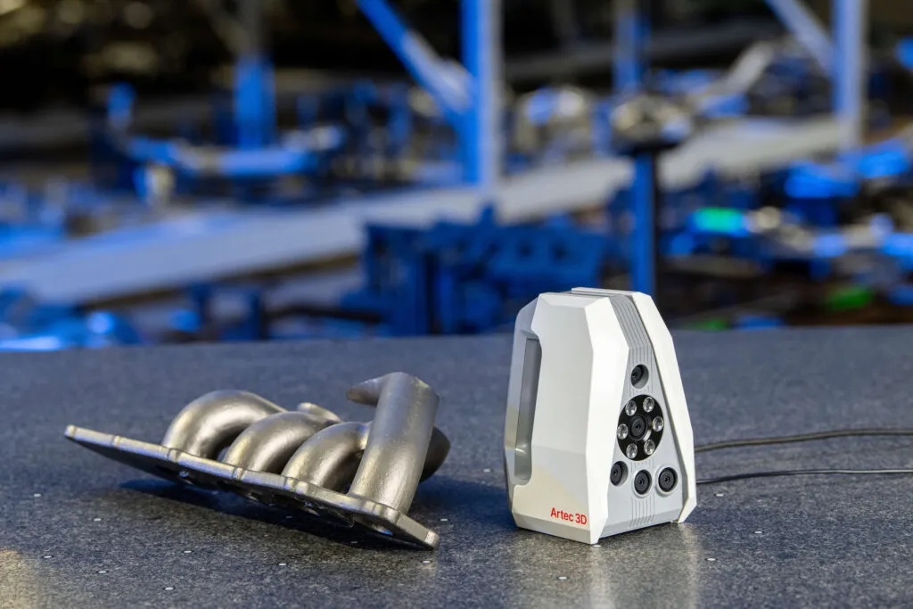 An Artec 3D scanner sitting on a workbench next to a metal automotive intake manifold, with a parts warehouse visible in the background.