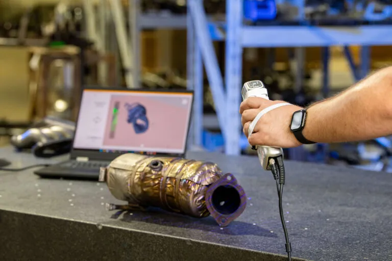 Handheld 3D scanner capturing a catalytic converter, with the scan shown on a laptop behind it