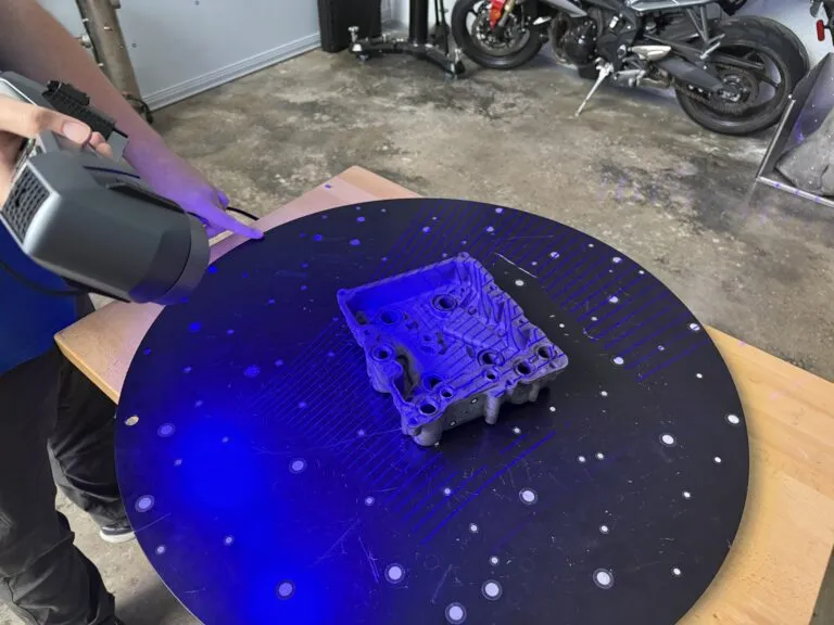 Handheld blue laser 3D scanner capturing a cast metal part on a marker-covered turntable