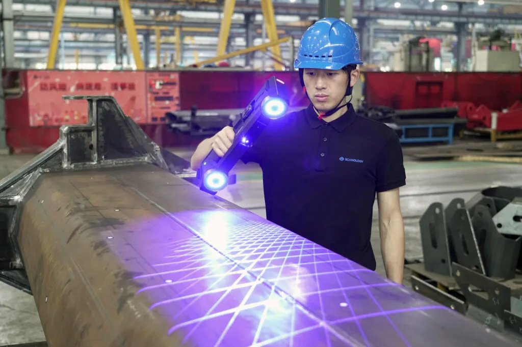 Operator scanning a large cylindrical steel weldment with the TrackScan GO handheld scanner projecting blue laser lines across the surface
